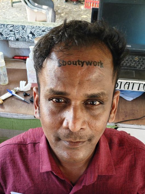 The image features a man with a tattoo of the word "bouttywork" written on his forehead, which appears to be a playful challenge or prank. The photo captures him looking directly at the camera, giving us an insight into his personality and sense of humor.