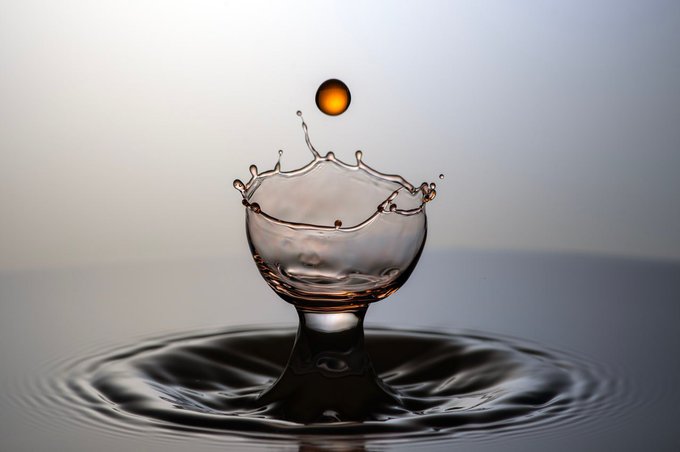A high-speed photograph captures a golden droplet suspended in the air above a splash forming a crown shape in a pool of water.