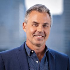 A professionally taken headshot shows a middle-aged man with short graying hair, wearing a dark blue blazer over a patterned blue button-down shirt, smiling warmly against a softly blurred office background. The image appears to be a portrait of Eric Balchunas, the Bloomberg analyst cited in the post about $IBIT and $GLD returning to the top 10 most-traded ETFs amid the shift from AI-focused semiconductor funds to a "debasement trade" narrative.