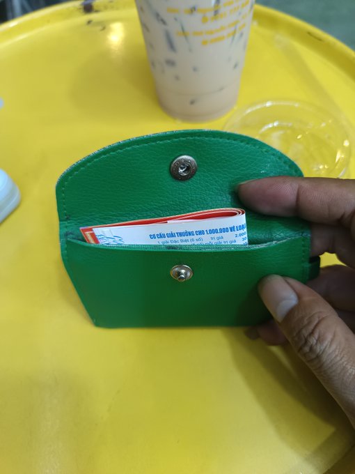 A pair of hands holds open a bright green leather wallet containing several Vietnamese Dong banknotes on a yellow table, next to a plastic cup of iced drink.