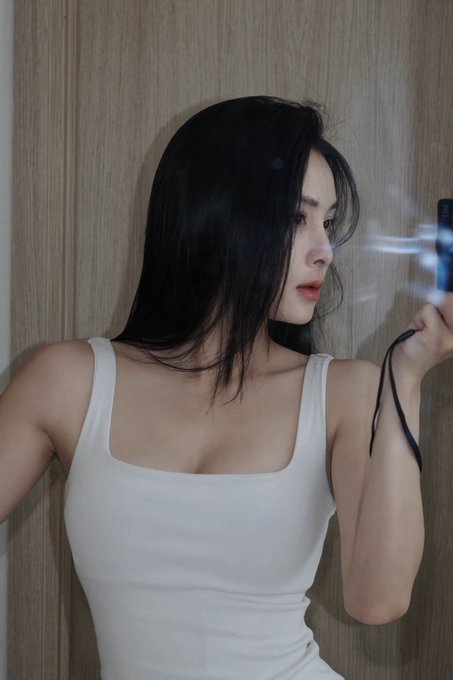 A young woman with long dark hair wearing a white tank top holds a camera, looking to the side against a wooden background.