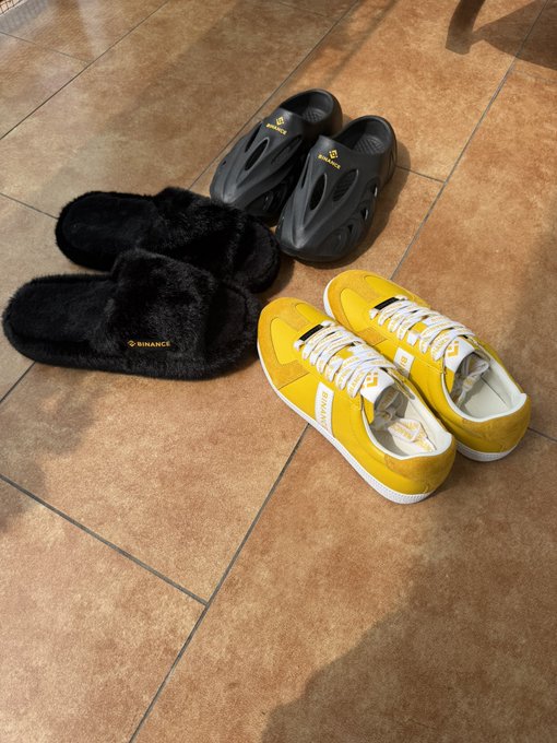 A collection of Binance-branded merchandise including black fuzzy slippers and gray slides, alongside a pair of yellow Nike sneakers, all arranged on a tiled floor as Mid-Autumn Festival gifts from Binance.