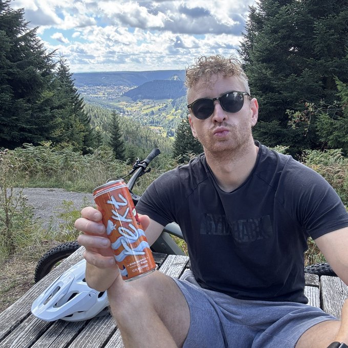 A man wearing sunglasses and a dark athletic shirt sits at a picnic table on a forested hillside, holding up an orange energy drink can toward the camera.