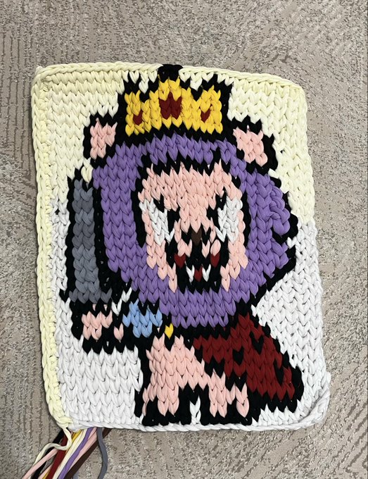 A knitted or crocheted tapestry depicting a pixel-art-style character with a crown, purple hair, and fangs, resting on a concrete surface.