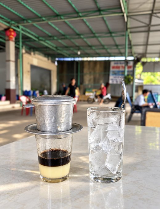 A traditional Vietnamese coffee filter (phin) dripping coffee over a glass of condensed milk, beside a tall glass of ice water, set in an open-air cafe.