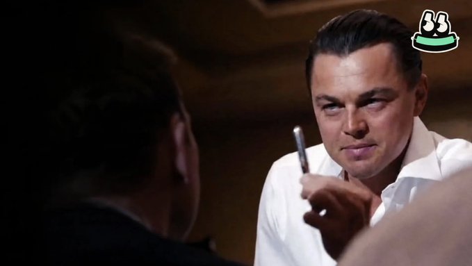 Leonardo DiCaprio, in his iconic role from "The Wolf of Wall Street," smirks while holding a pen, perfectly mirroring the post's "Sell me the pen" challenge—a legendary scene symbolizing persuasion and salesmanship used here as a metaphor for pitching the next penny stock pick.