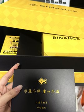 The image shows a person holding up a small black box with gold lettering that says "Binance" on it, along with some text written in Chinese characters. The box appears to be a gift from Binance, possibly containing a digital currency or other cryptocurrency-related items.