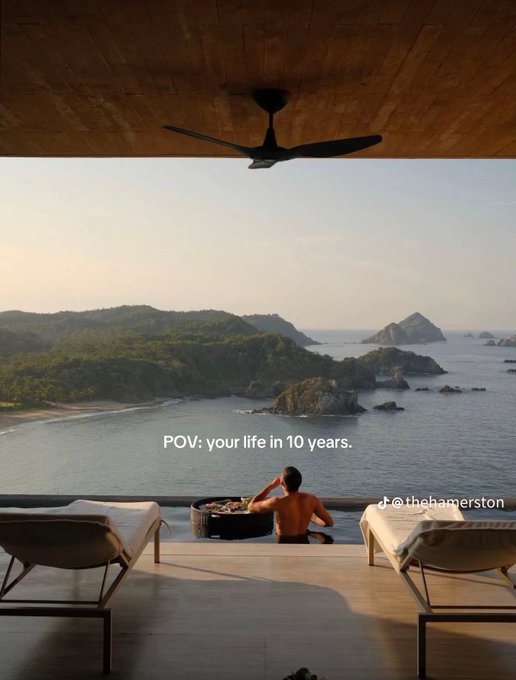 A POV-style image looking out from a luxurious open-air room with sun loungers and a ceiling fan, showing a person sitting by a hot tub facing a scenic coastline with islands and mountains, overlaid with the text 'POV: your life in 10 years.' and '@thehamerston'.