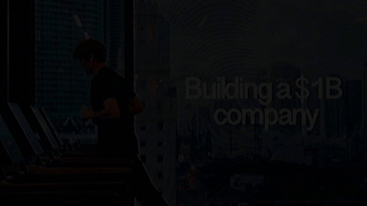 The image shows a man running on an exercise machine while wearing headphones. The text "Building a $1B company" is displayed above him, indicating that he might be working for a company related to fitness or health. The background of the image features a city skyline with tall buildings and a window in one of them.
