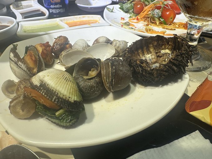 A white plate filled with various cooked shellfish, including clams and a large crustacean, sits on a table alongside condiments, a Pepsi bottle with Vietnamese text 'NGON GHIỀN PEPSI', and a side salad.