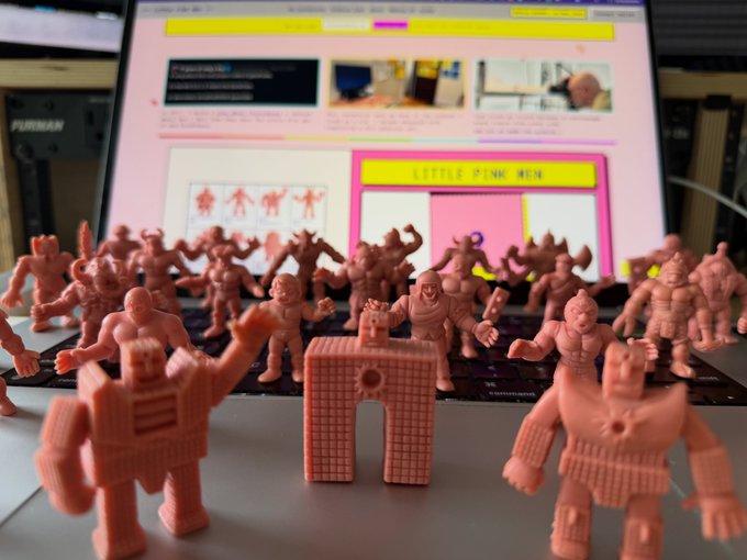A collection of pink/flesh-colored 3D-printed monster and robot action figures sits on a desk in front of a computer screen displaying a design website.