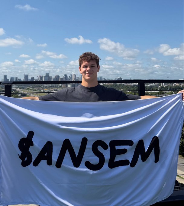 A young man stands on a rooftop balcony against a city skyline backdrop, smiling as he holds up a large white flag displaying "$ANSEM" in bold black lettering, carrying out the promised in-real-life $ANSEM airdrop.