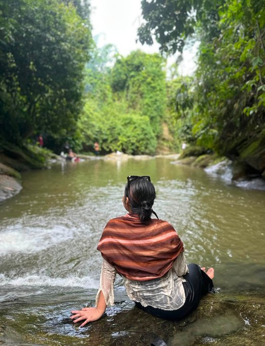 A person wearing a striped shawl and sunglasses on their head sits in a shallow stream surrounded by lush green jungle vegetation, looking out at other people in the distance.
