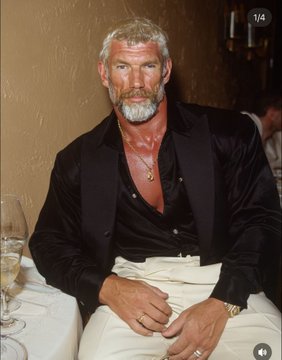 An older man with a gray beard and mustache, dressed in a black satin shirt and cream pants, sits at a table in what appears to be a restaurant setting.
