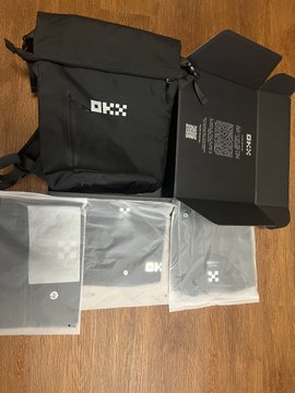 Unboxing photo of OKX branded merchandise including a black backpack, a black box, and clothing items in plastic packaging on a wooden floor.