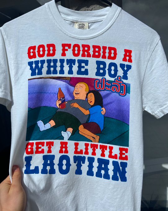 A white t-shirt displays the text 'GOD FORBID A WHITE BOY GET A LITTLE LAOTIAN' in red and blue lettering, surrounding an illustration of the King of the Hill characters Bobby and Connie, with Laotian script included.