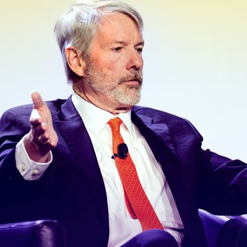 A photograph of a man with a beard wearing a dark suit and red tie, gesturing with his hand while speaking at what appears to be a panel discussion or conference.
