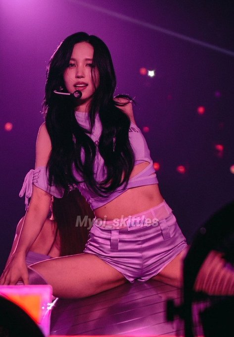 A woman with long dark hair, wearing a lavender cut-out top and matching shorts along with a headset microphone, is kneeling on stage under purple concert lighting. A watermark reading "Myoi_skittles" is visible across the image.