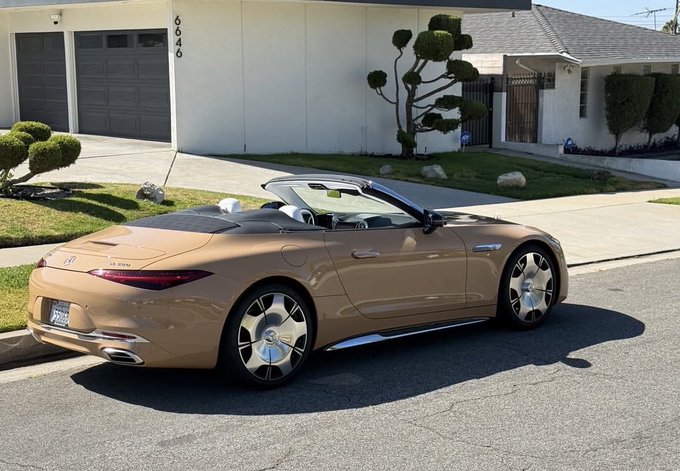 A tan/gold-colored Mercedes-Benz SL convertible parked on a suburban street in front of a house with decorative topiary trees.