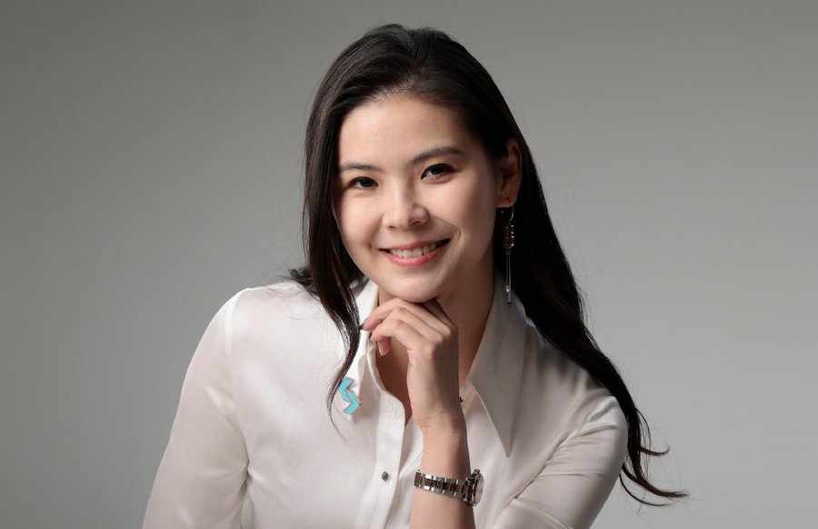 A professional headshot of a smiling Asian woman with long dark hair, wearing a white satin blouse and a wristwatch, posing against a gray background.