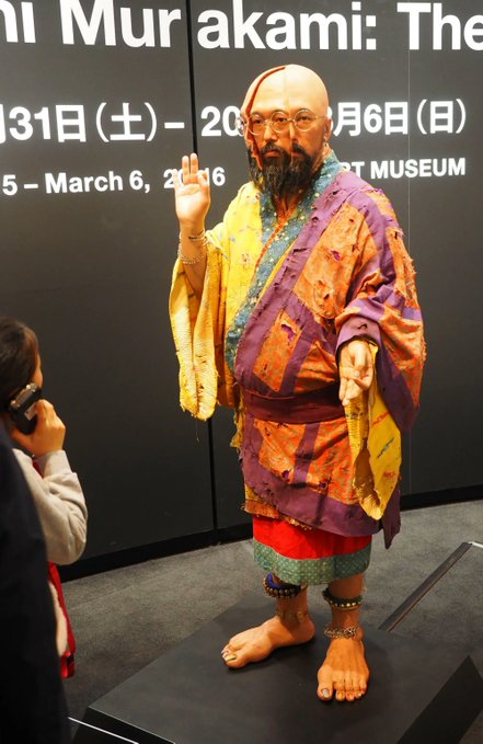 A realistic sculpture of the Buddhist monk Musō Sakyō by artist Etsuro Murakami, exhibited at a museum from 2015–2016, depicting the bearded figure in colorful patchwork robes with one hand raised.