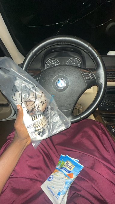 A person sits in the driver's seat of a BMW holding a clear plastic bag labeled 'Bubble Tea By Dimple,' with a pack of Great Value Salted whipped cream resting on their maroon lap.