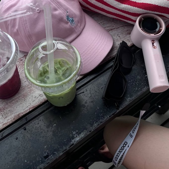 A tabletop scene showing a pink Ralph Lauren cap, a green matcha drink with a straw, sunglasses, a small pink portable fan, and a wristband.