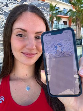 The image features a woman holding up her phone, which displays an image of a pizza with the text "Free pizza hits different" written below it. The woman appears to be smiling as she shows off the photo. In the background, there is a building visible behind the woman and another person standing further back in the scene.