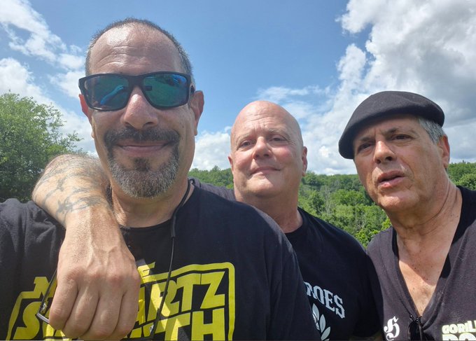 A selfie of three middle-aged men outdoors under a partly cloudy sky; the man on the left wears a Metallica t-shirt and blue reflective sunglasses, the center man wears a black t-shirt, and the right man wears a black flat cap.