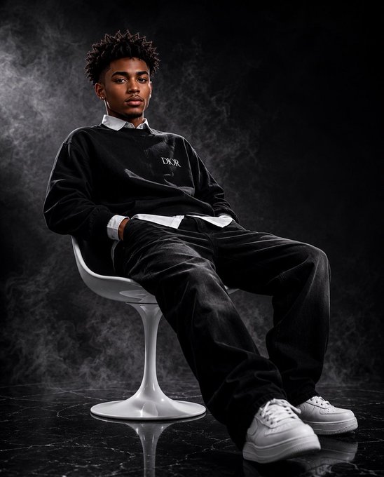 A young man with short textured hair sits on a white modern chair against a dark, smoky background, wearing a black Dior sweater, black pants, and white sneakers.