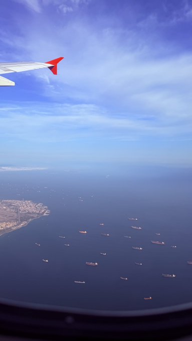 An aerial view from an airplane window showing the wing and numerous cargo ships scattered across a vast blue ocean.