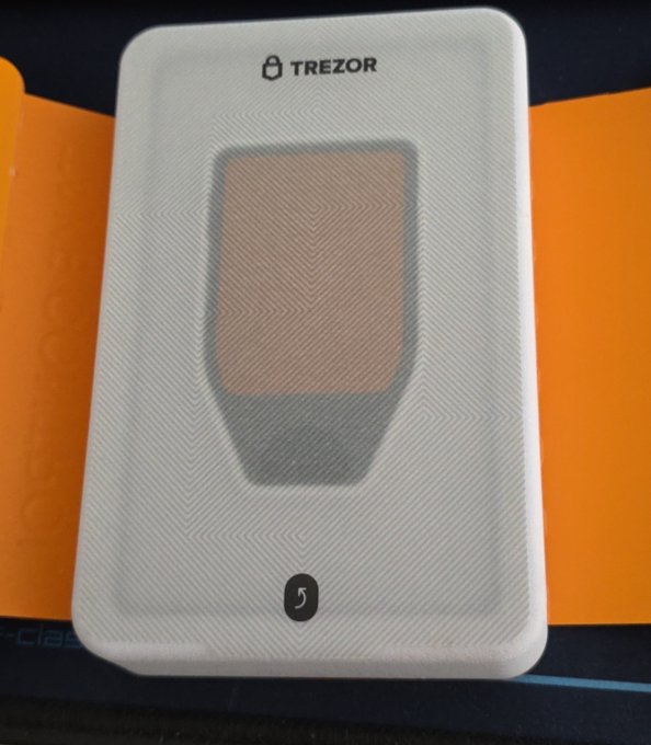 A white Trezor hardware wallet device with a copper/gold colored touchscreen display and a return arrow button, photographed against an orange background.