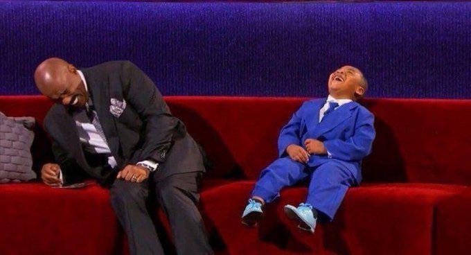 A man in a dark suit and a young boy in a blue suit sit together on a red couch, both laughing joyfully during what appears to be a television appearance.