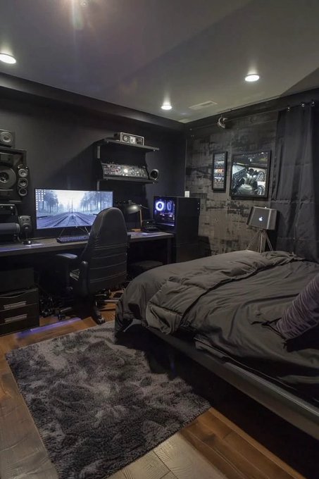 A dimly lit, moody bedroom featuring a gaming/streaming setup with dual monitors displaying a black-and-white train scene, studio monitors, and a gaming chair beside a neatly made bed, with framed posters on a dark wall. The image visually echoes the caption's theme of a late-night grind—trading screens glowing in the darkness as the creator signs off with a motivational "goodnight" message to their followers.