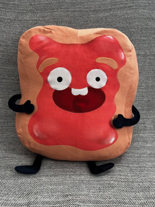 A plush pillow shaped like a slice of peanut butter toast with jelly, featuring a smiling face, googly eyes, and small black arms and legs, sitting on a gray textured surface.