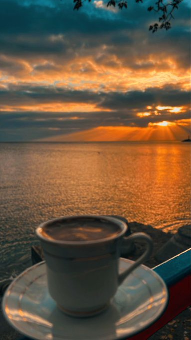 A small cup of coffee sits on a saucer in the foreground, with a dramatic sunset over a calm body of water in the background.