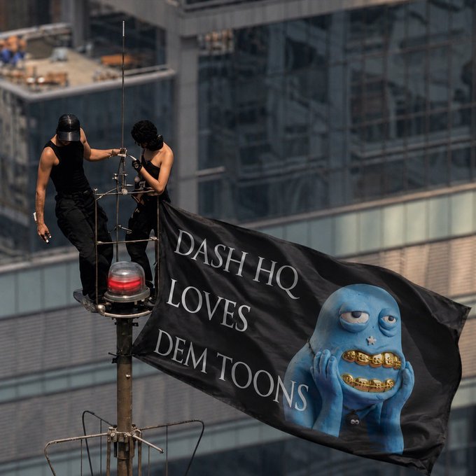 Two people dressed in black stand on a high-rise antenna platform unfurling a large black flag that reads "DASH HQ LOVES DEM TOONS" alongside a blue cartoon character with gold grills. The stunt playfully mirrors the post's announcement of surprising fans in their DMs, referencing "Angelina & Ivan" as they take the @just_t00ns mascot to new heights—literally.