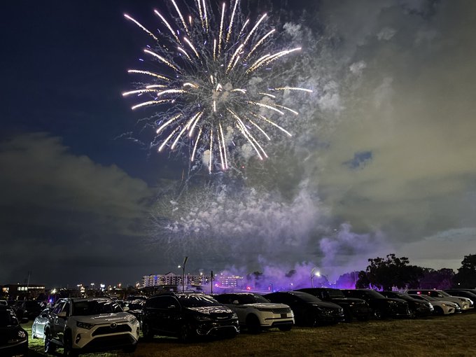 A large fireworks display explodes in the night sky over a parking lot full of cars.