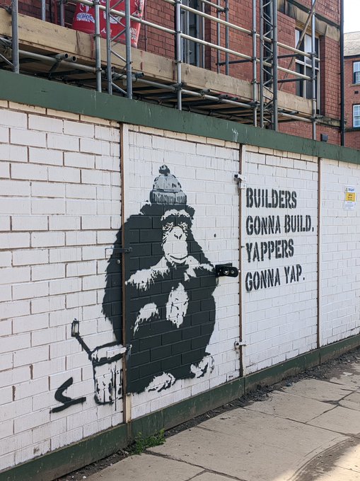A black and white street art mural on a white brick wall depicting a chimpanzee wearing a beanie and painting with a brush, accompanied by the text 'BUILDERS GONNA BUILD, YAPPERS GONNA YAP.'