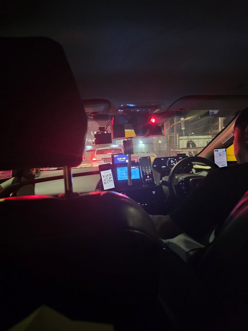 A nighttime view from the back seat of a car shows a driver, illuminated dashboard screens with navigation, a mounted phone displaying a QR code, and red traffic lights visible through the windshield.