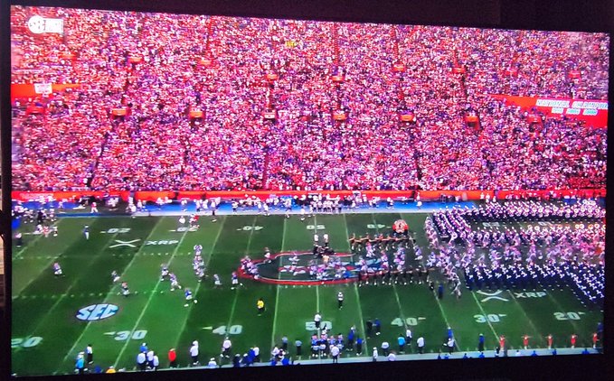 A packed football stadium at the Super Bowl XLI (XLI logo visible on the field and "Super Bowl" signage), with players lined up on the field and a marching band assembled at midfield during pregame. The post "Gametime. X. R. P. 🔥" celebrates the big-game atmosphere shown in the image, with the Roman numeral "XLI" from the field matching the energy of the kickoff moment.