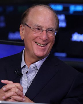 A smiling bald man with glasses in a dark blazer sits with his hands folded, set against a backdrop of digital screens—appearing to be BlackRock CEO Larry Fink, whose firm recently purchased $138.9 million in Bitcoin, signaling the return of institutional crypto investment.