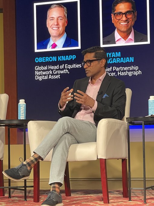 The image features a man sitting in a chair, talking at a conference or event. He appears to be wearing glasses and a suit, suggesting that he might be a speaker or an expert in his field. The background of the image shows two other people present as well, possibly fellow speakers or attendees. There are also chairs visible in the scene, indicating that this is likely a formal setting for a discussion or presentation.