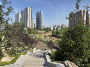 The image shows a cityscape with tall buildings, including skyscrapers, lining up along a street. The perspective of the photo appears to be taken from a higher vantage point, providing an overview of the urban environment. In the foreground, there is a stone staircase leading down into a park area filled with trees and greenery. The post accompanying this image provides information about the location and possibly some historical context related to the city's development or landmarks in the area.