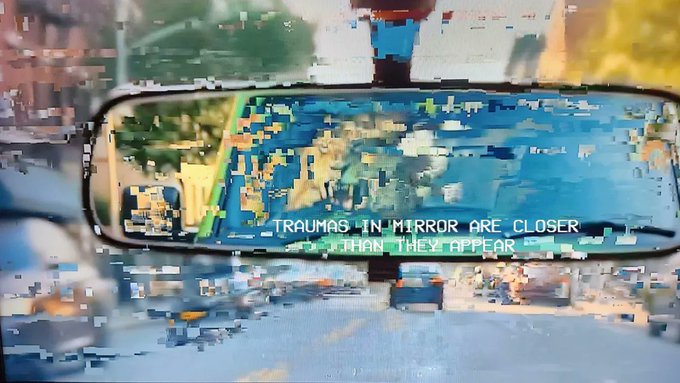 Glitchy, pixelated image of a rearview mirror reflecting a busy city street with the text 'TRAUMAS IN MIRROR ARE CLOSER THAN THEY APPEAR' displayed across the reflection.