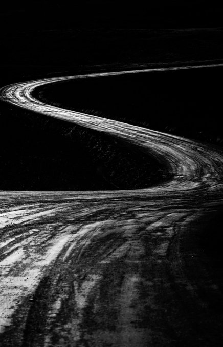 A high-contrast black and white image of a winding road or river path with textured tire tracks or ripples, curving through a dark, shadowy landscape.