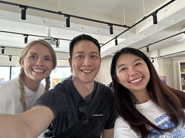 Three smiling people pose for a selfie indoors at what appears to be a modern office or co-working environment with bright lighting.