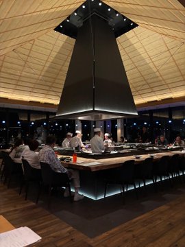 Interior of an upscale Japanese restaurant featuring a large central hooded grill, a curved counter with seated diners, and modern wooden ceiling architecture.