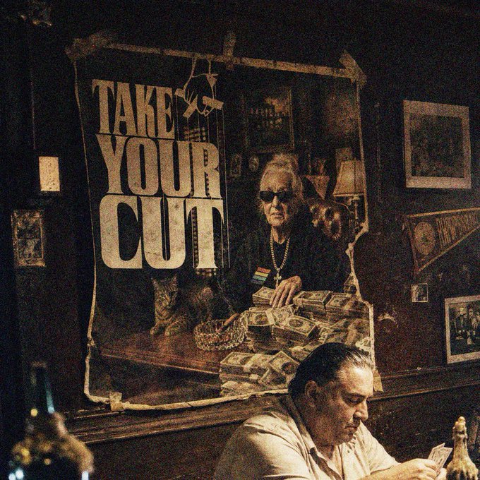 Photo of a dimly lit bar interior featuring a vintage 'TAKE YOUR CUT' poster with an illustration of an elderly woman surrounded by stacks of money, with a bartender working in the foreground.