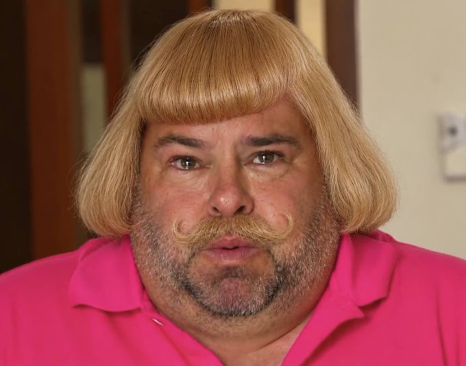 A close-up portrait of a man with a bowl-cut blonde wig and curled mustache, wearing a bright pink polo shirt.